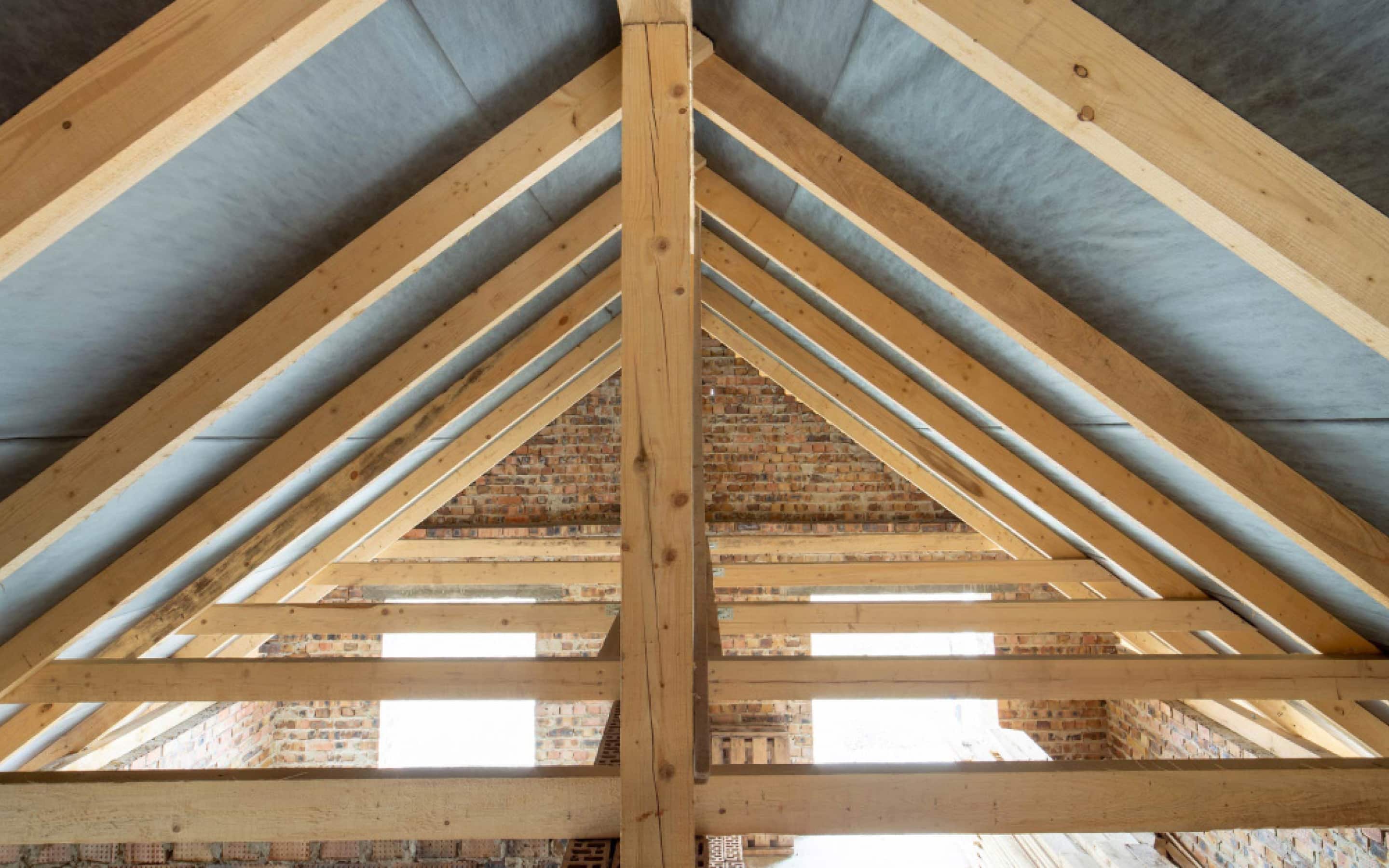 Interior view of the wooden rafter and truss structure of a gable roof under construction, with a waterproof membrane and brick walls in the background.