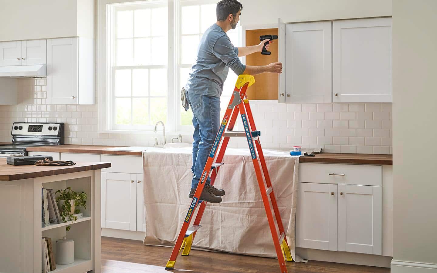 An individual uses a cordless drill on an orange step ladder in a kitchen. 