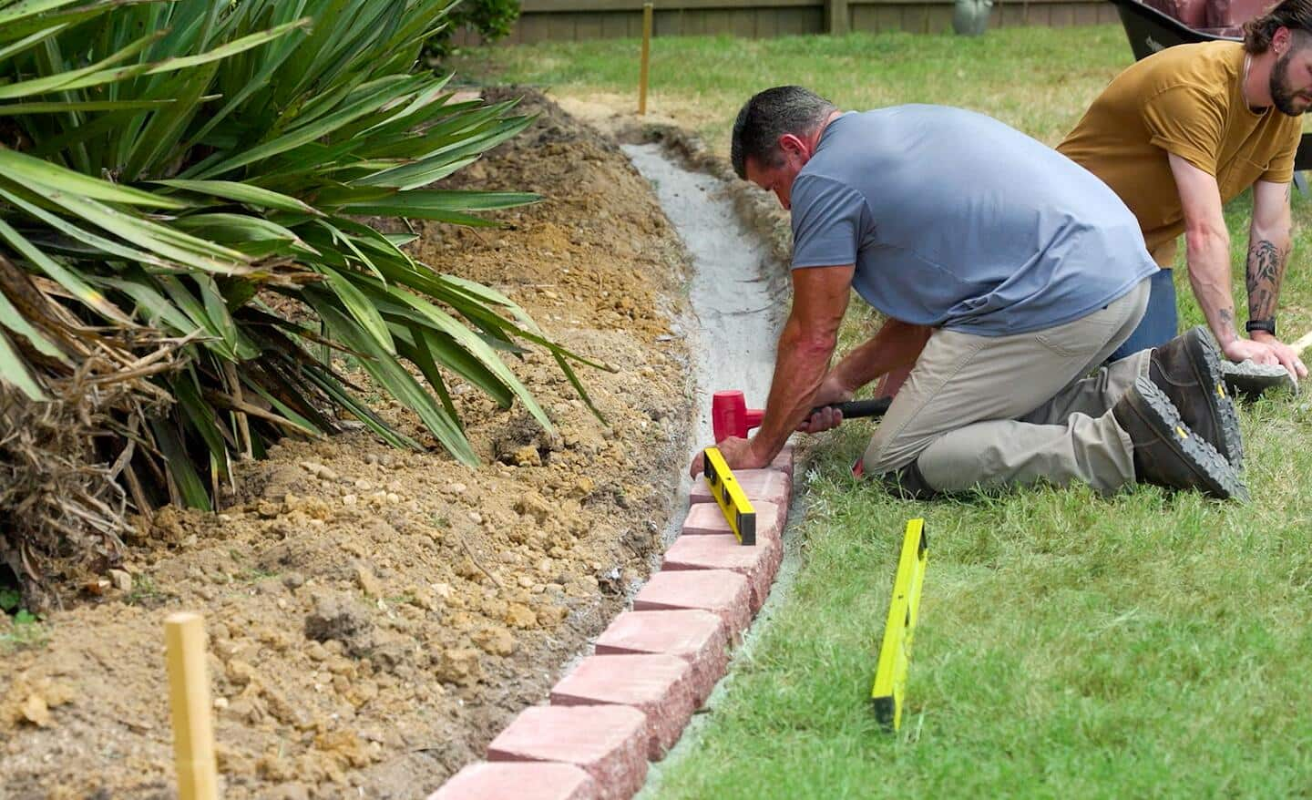 A man places a retaining wall block and levels it with a mallet while another man readies the next block.