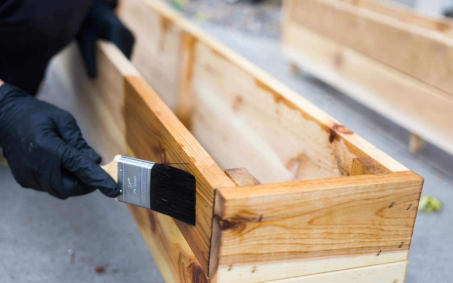 A gloved person uses a black paintbrush to apply stain to a window box.