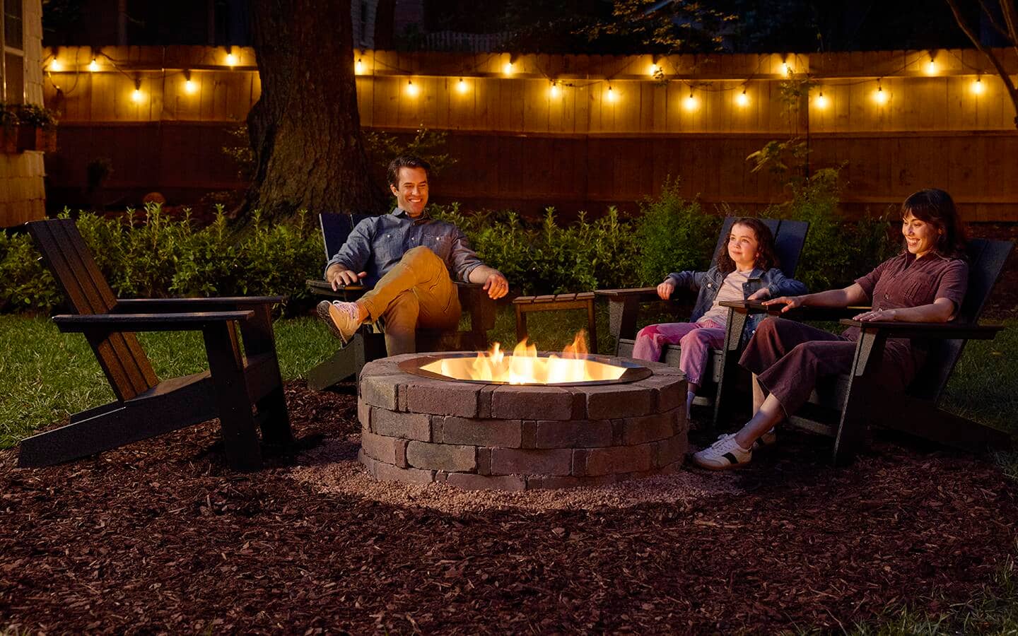 A family sitting in Adirondack chairs around a stone fire pit with electric lights in the background.
