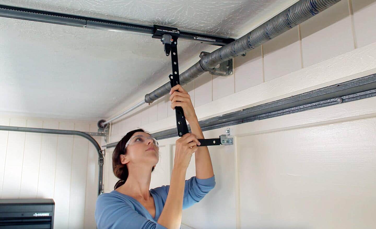A woman wearing safety goggles connects the power unit of a garage door opener to the ceiling. A woman wearing safety goggles connects the power unit of a garage door opener to the ceiling.