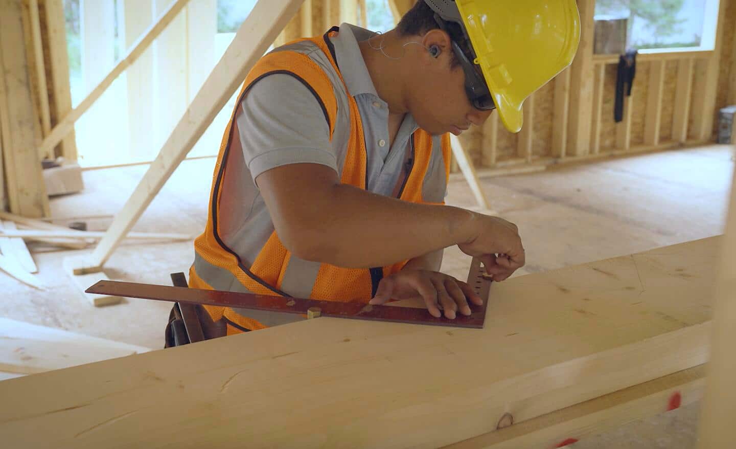 A construction worker uses a framing square with stair gauges and a pencil to mark stair stringer cut lines on a 2x12 piece of lumber.
