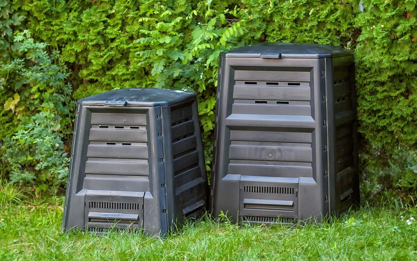 Two gray plastic compost bins placed outdoors in front of a hedge.