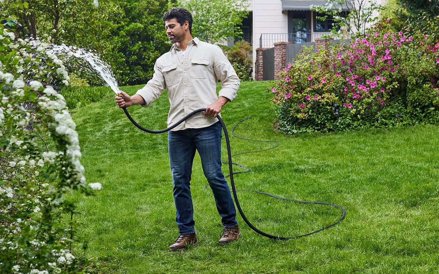 An individual holding a black garden hose sprays water onto a shrub in a yard.
