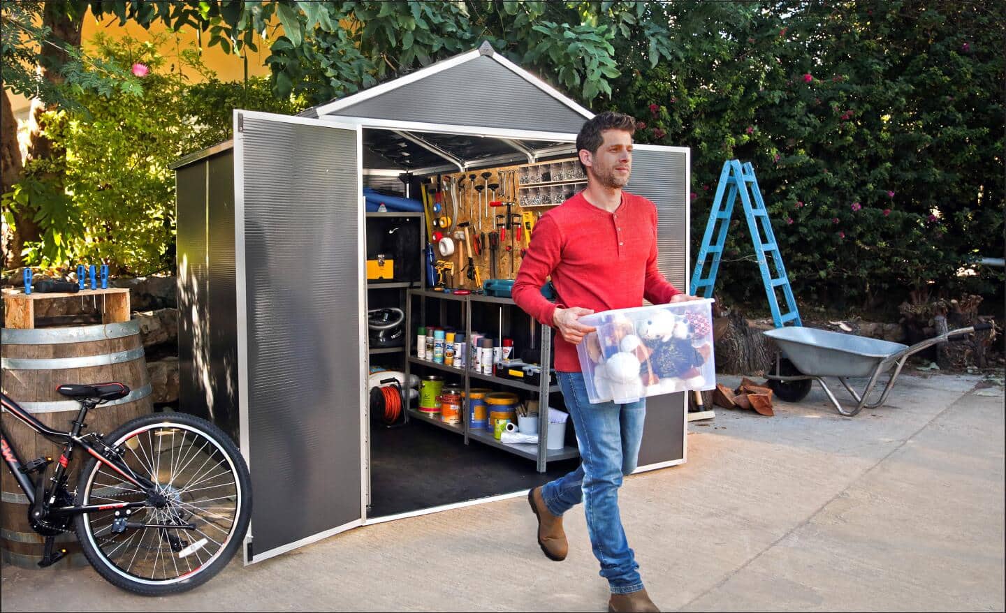 A man carries a clear plastic tote outside of a small garden shed.