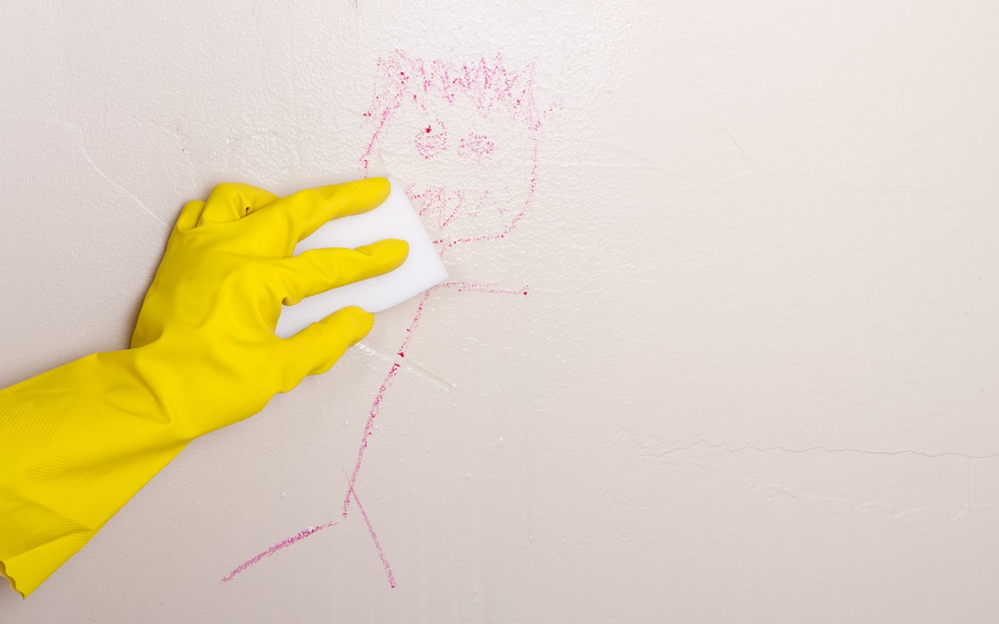 A person wearing gloves uses an erasing pad to clean a crayon drawing from a wall. A person wearing gloves uses an erasing pad to clean a crayon drawing from a wall.