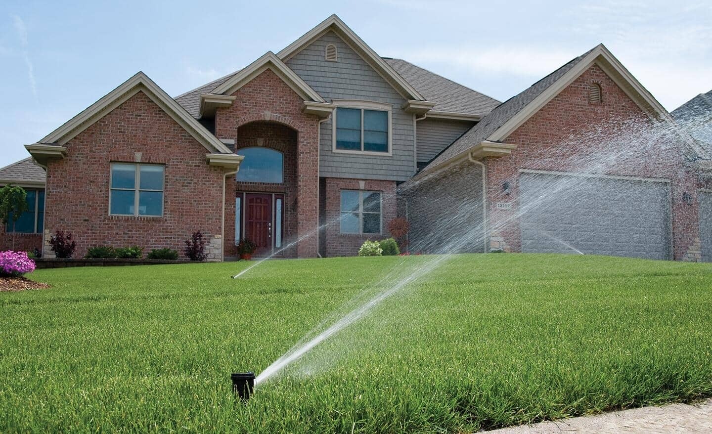 Sprinklers water a green lawn in front of a house.