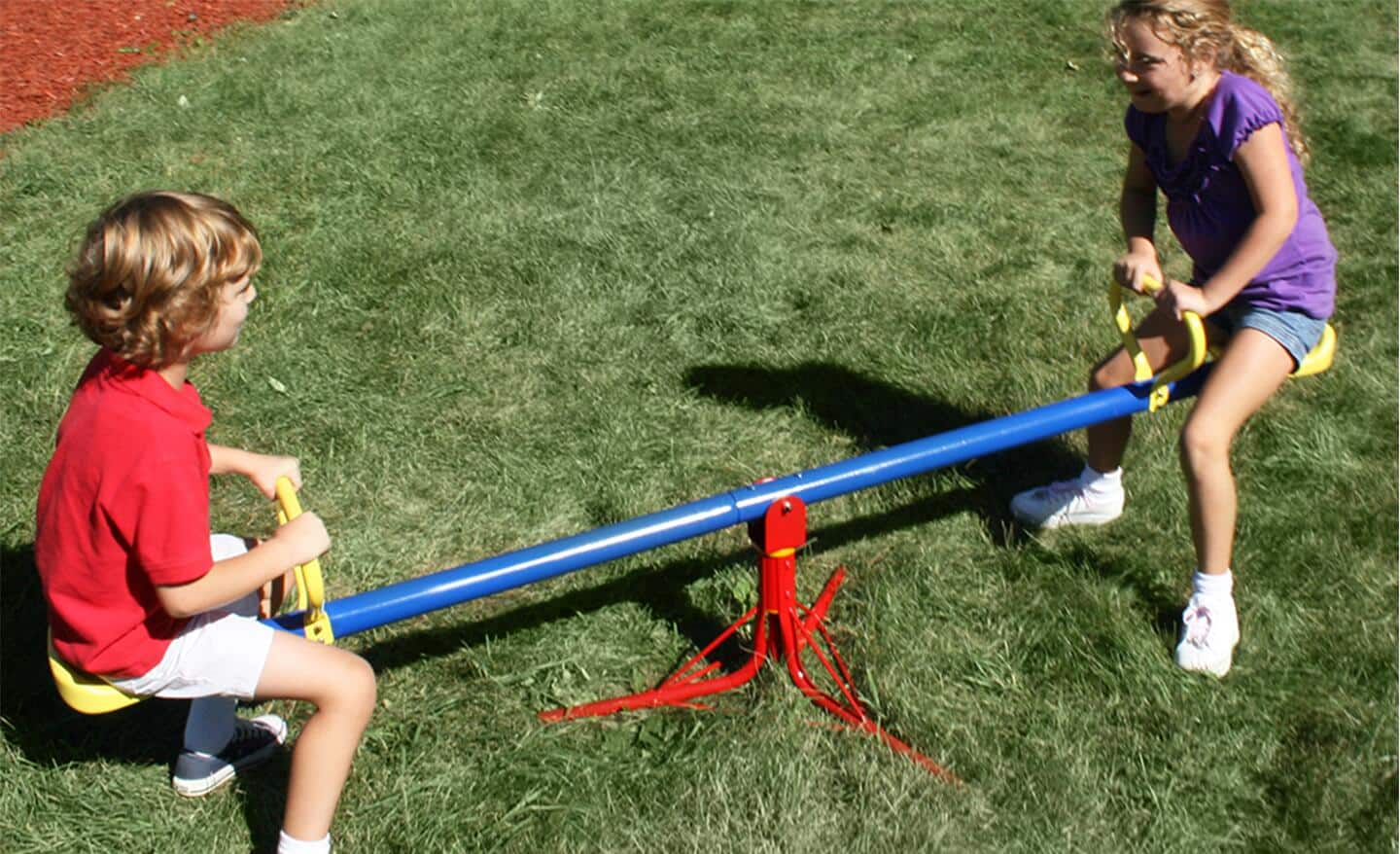 A pair of children playing on a blue, yellow and red playground seesaw.