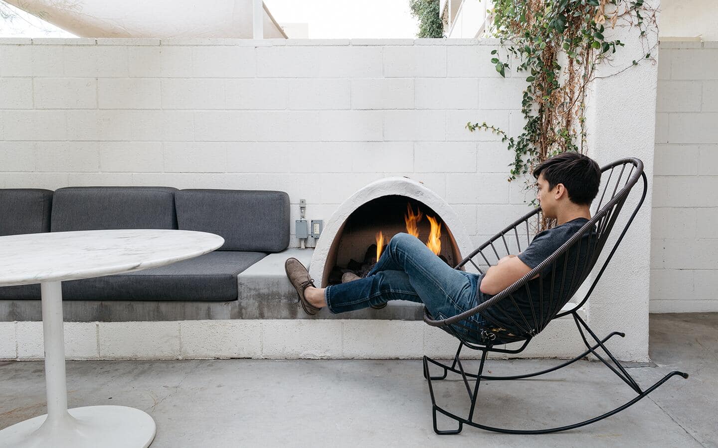 A man in a metal egg chair relaxing by a modern outdoor fireplace.