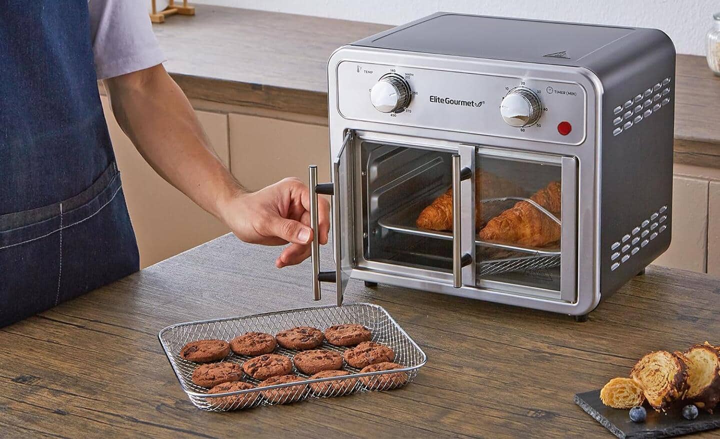 A stainless steel air fryer with double French doors sits on a counter. It has croissants inside and tray of cookies next to it. A person is holding one of its door handles.