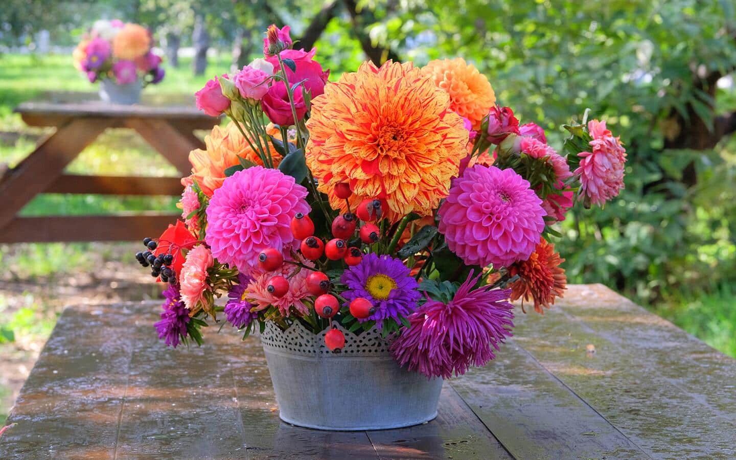 A silver bucket full of colorful cut flowers sitting on a picnic table.
