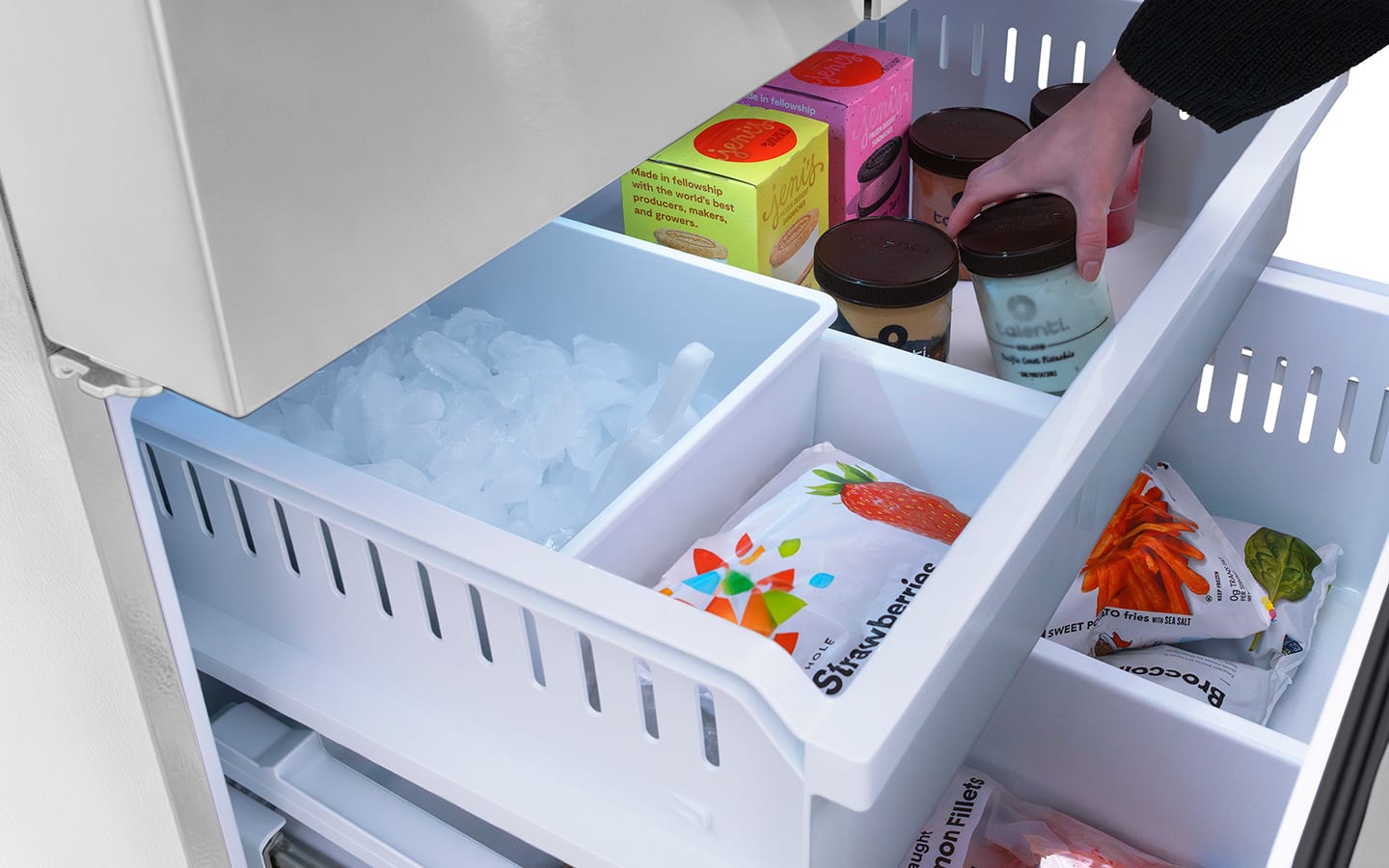 A person reaches into an open sliding drawer in the bottom half of an upright freezer.