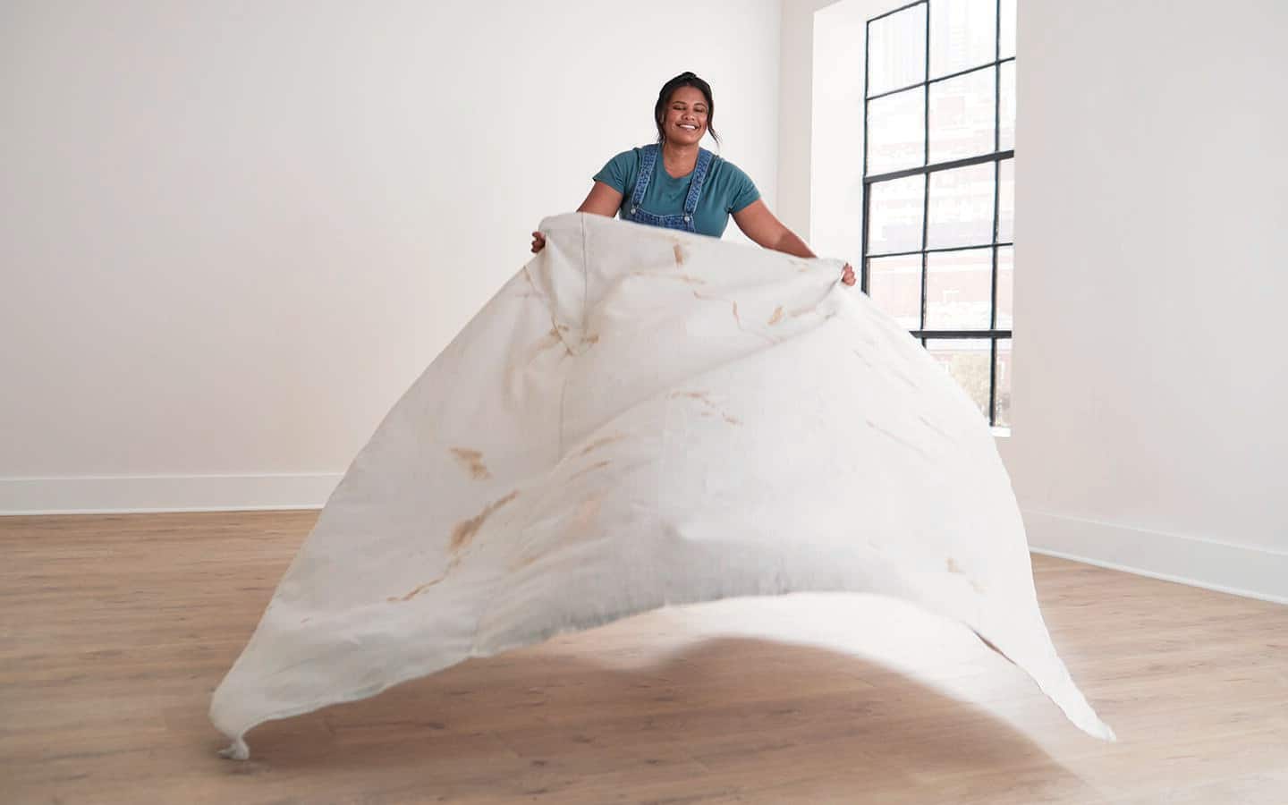 A woman lays a drop cloth on a wood floor.