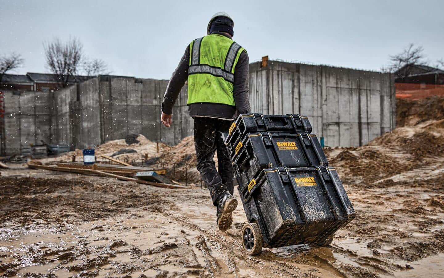 A construction worker pulls a modular tool storage system through a muddy job site with a concrete wall in the background.