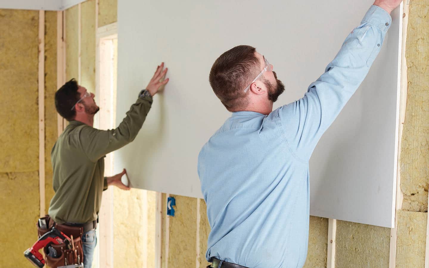 Two people in safety glasses hang a piece of drywall in a room that's under construction. Two people in safety glasses hang a piece of drywall in a room that's under construction.