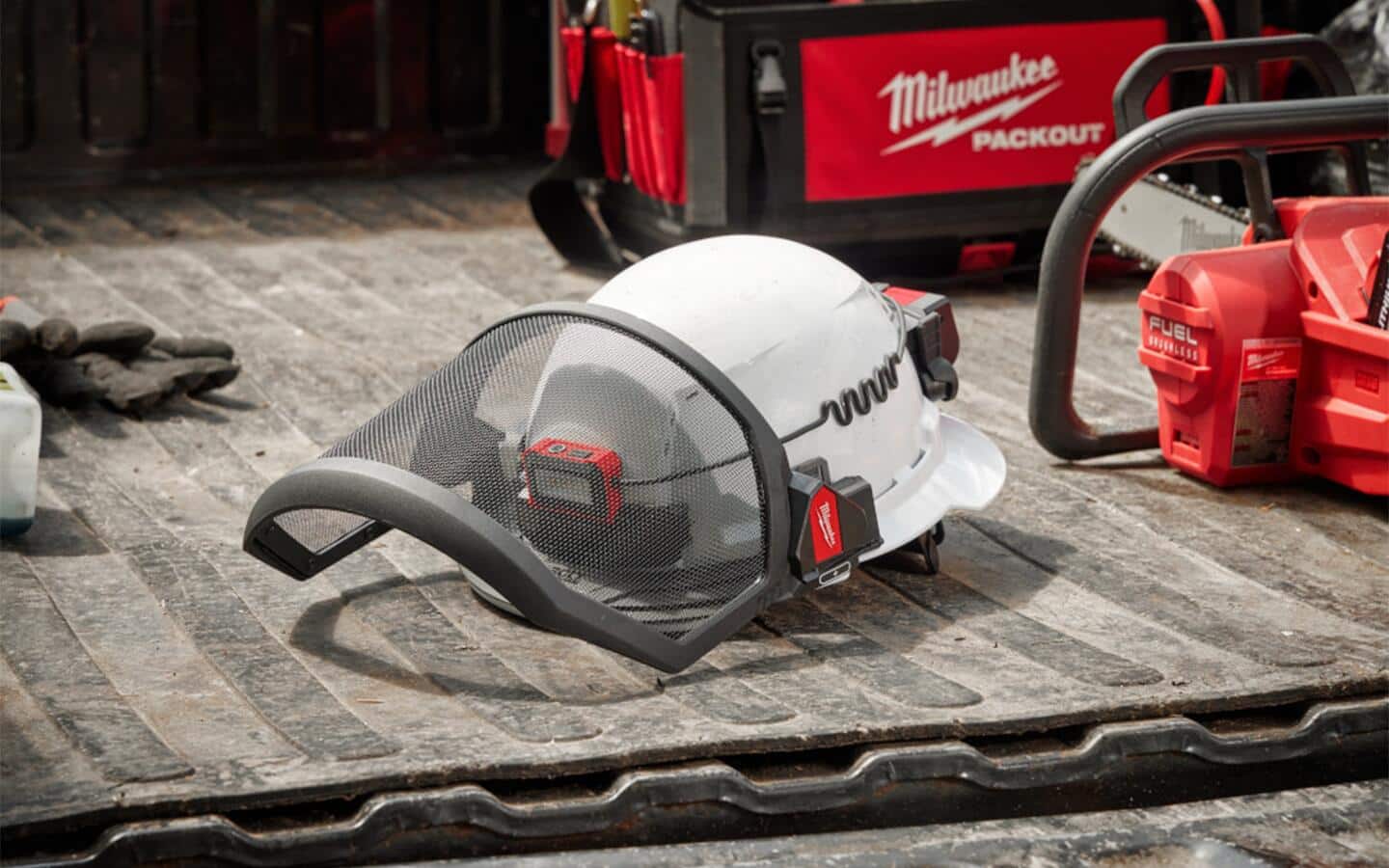 A hardhat with a light and mesh face shield sits in the back of a truck with a chainsaw and toolbox visible behind it. A hardhat with a light and mesh face shield sits in the back of a truck with a chainsaw and toolbox visible behind it.