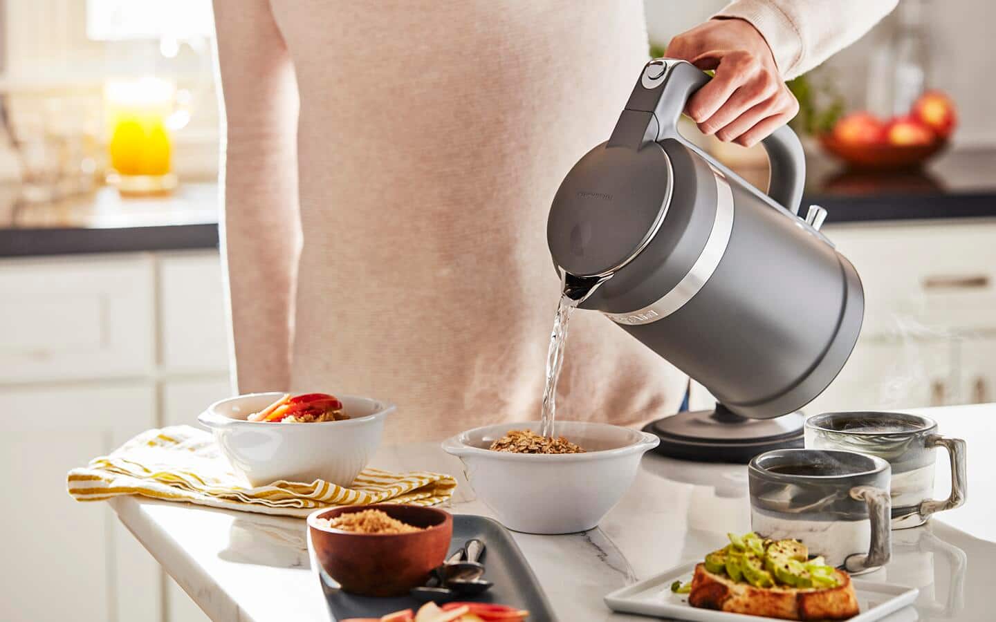 A person uses an electric kettle to pour water into a bowl of oatmeal.