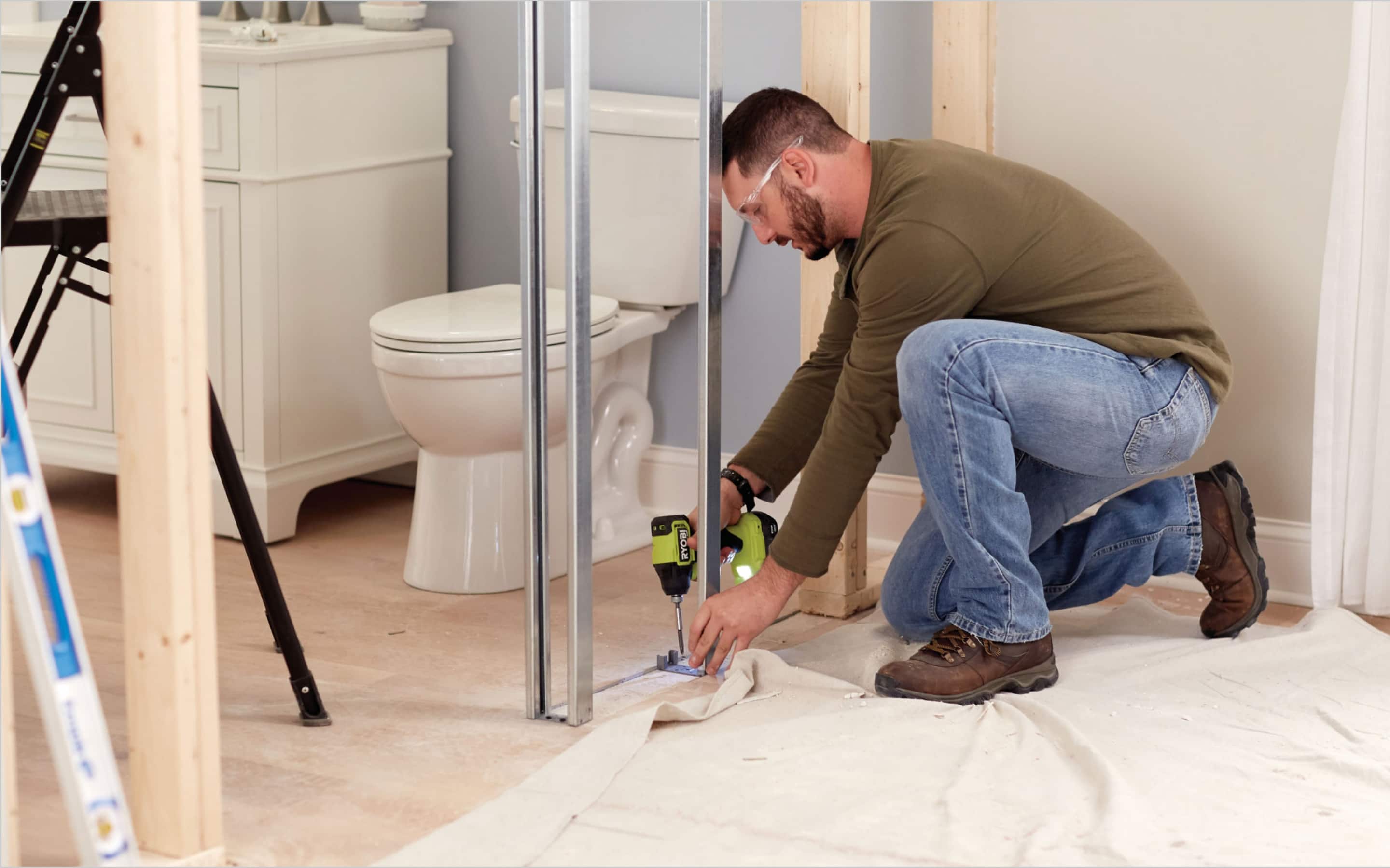 Un contratista atornilla un perfil metálico al piso durante la remodelación de un baño con inodoro y vanidad al fondo.