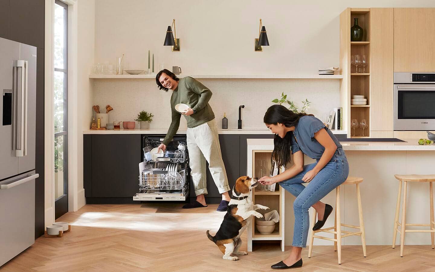 A man empties a dishwasher in a smart kitchen, while a woman feeds a dog. A man empties a dishwasher in a smart kitchen, while a woman feeds a dog.