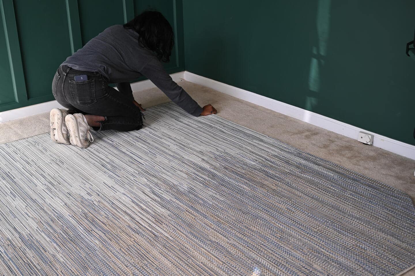 Lauren is laying out the area rug that covers the office floor. The rug is a striped woven mix of neutral colors and a deep blue. 