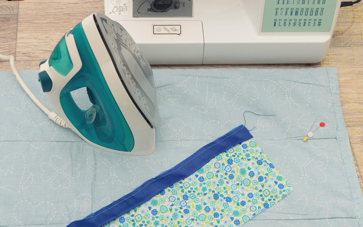 A green-and-white dry iron on a table next to a sewing machine. A green-and-white dry iron on a table next to a sewing machine.