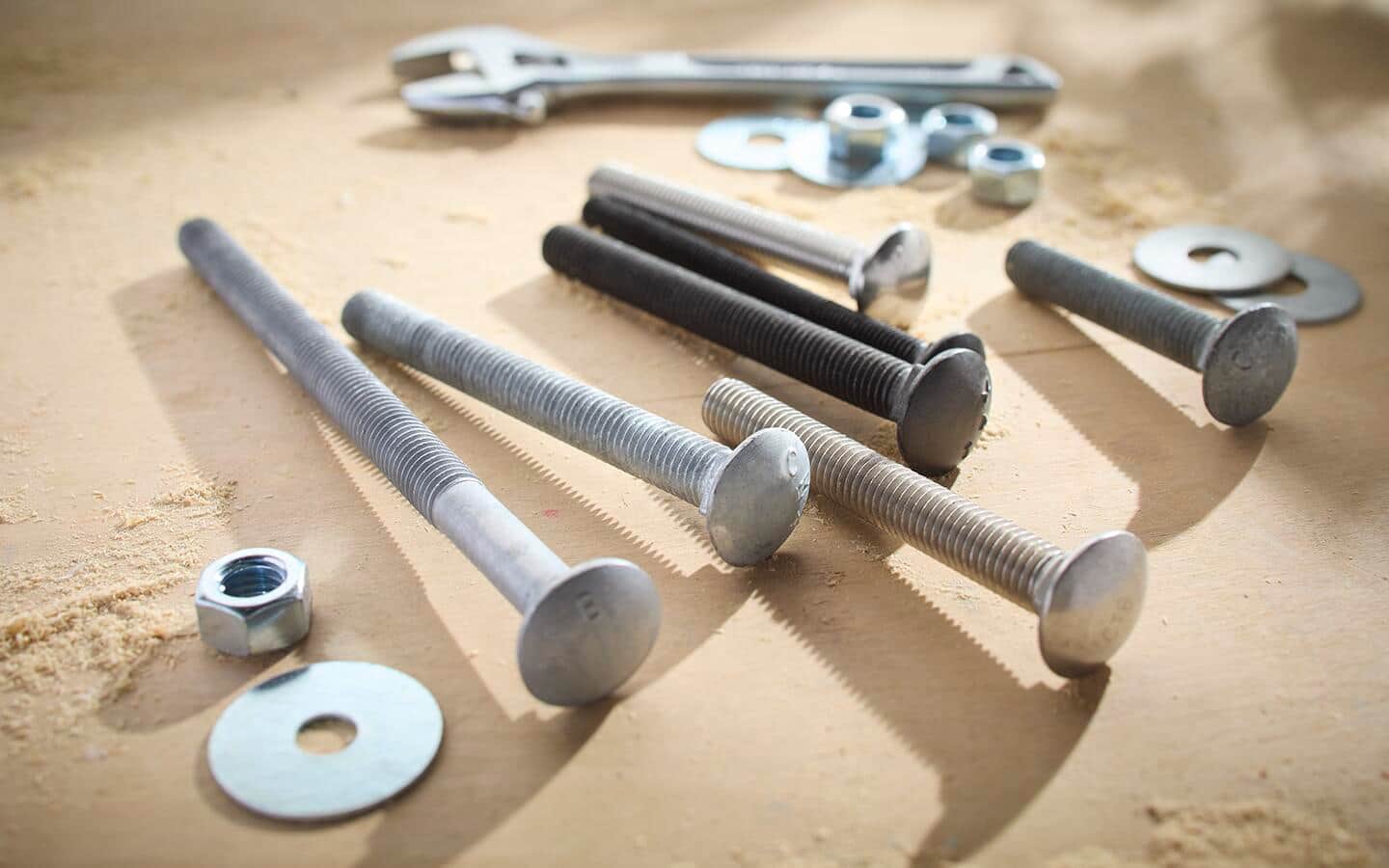 Carriage bolts in several finishes and sizes among an assortment of nuts and washers with an adjustable wrench in the background.