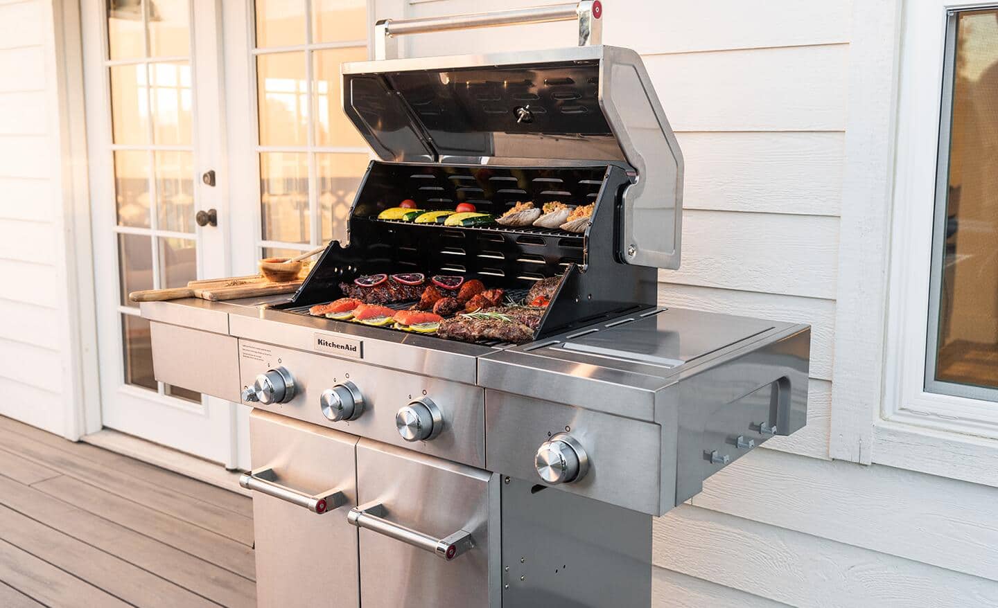 Meat and vegetables cook on a gas grill that stands on a deck.