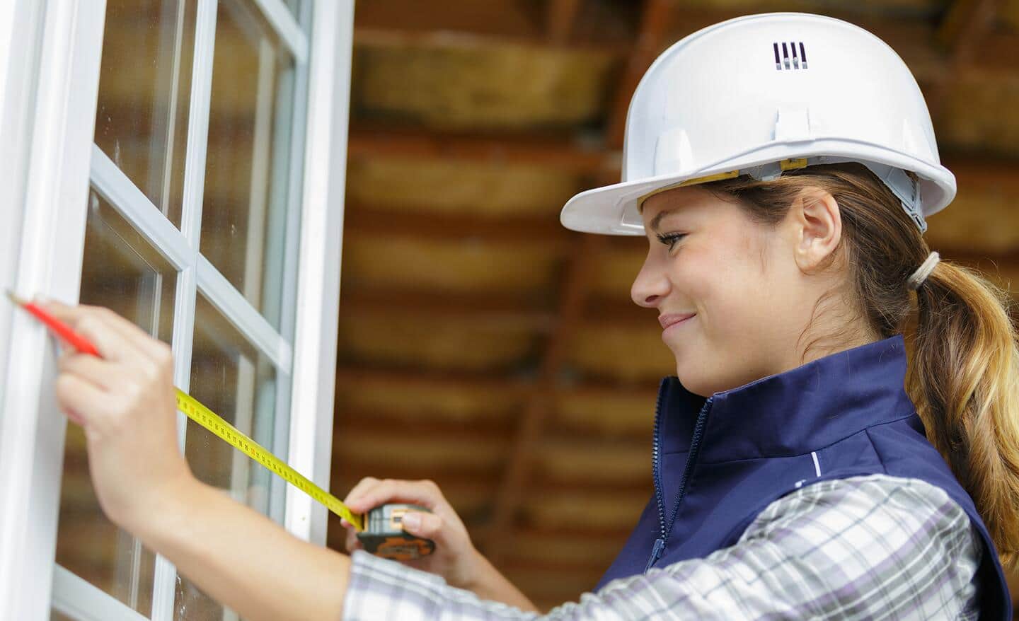 A woman wearing a hardhat measures a window with a tape measure.
